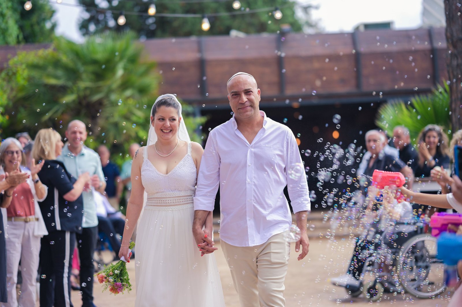 החתן והכלה צועדים יד ביד בין האורחים ובועות סבון באוויר